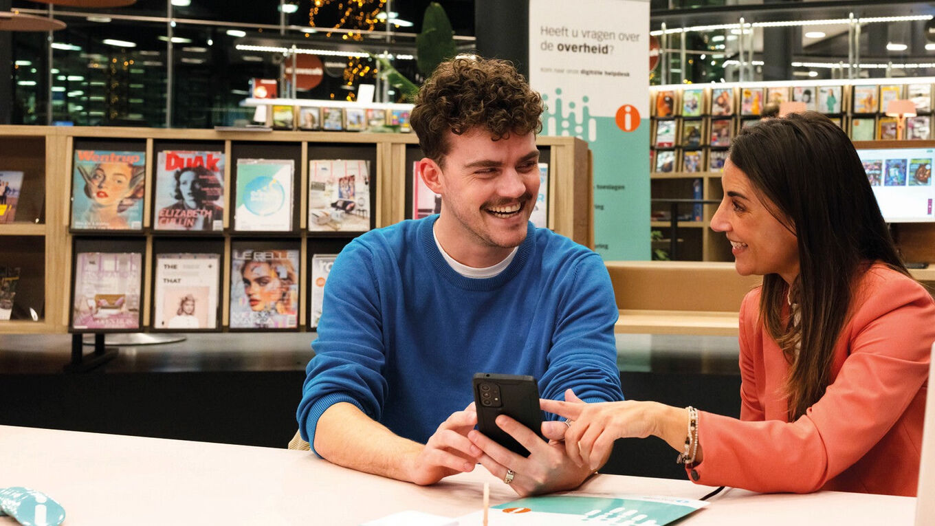 Jonge man met hip snorretje en telefoon vraagt om toelichting aan een medewerker van het IDO